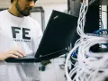 Inżynier w koszulce "FE FIELD ENGINEER" sprawdza sieć na laptopie. Analizuje połączenia, jakby sprawdzał, tracert co to jest.