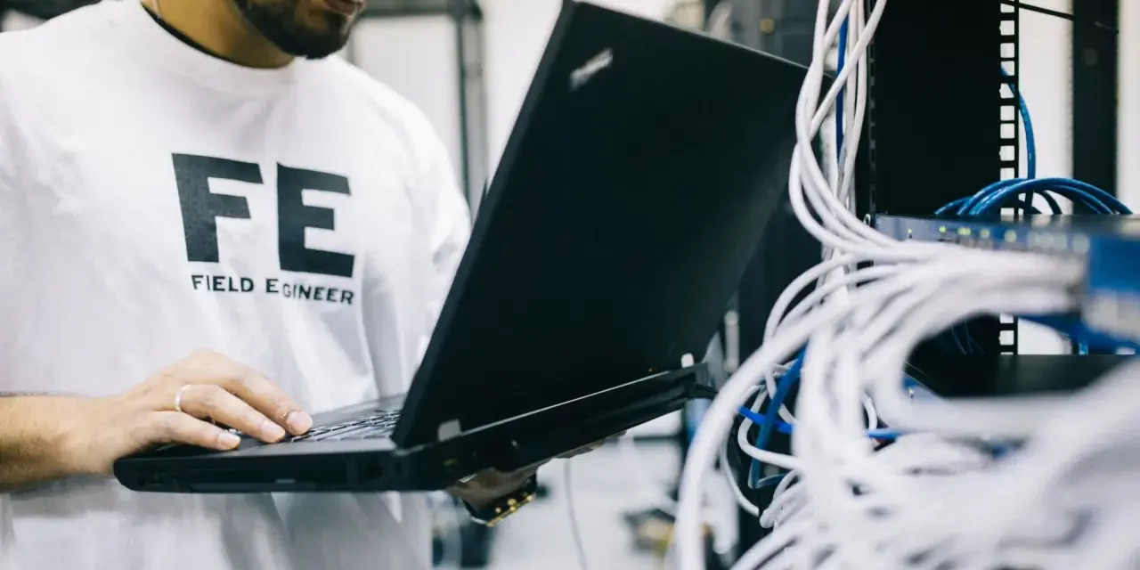 Inżynier w koszulce "FE FIELD ENGINEER" sprawdza sieć na laptopie. Analizuje połączenia, jakby sprawdzał, tracert co to jest.