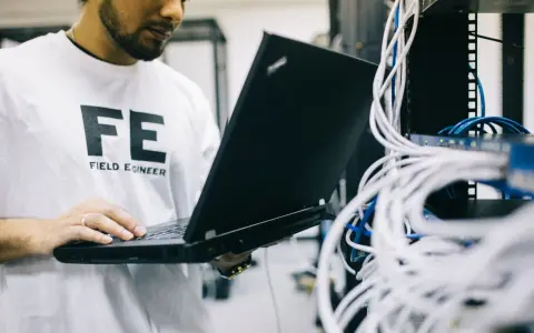 Inżynier w koszulce "FE FIELD ENGINEER" sprawdza sieć na laptopie. Analizuje połączenia, jakby sprawdzał, tracert co to jest.