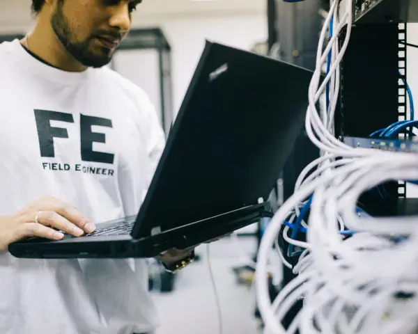 Inżynier w koszulce "FE FIELD ENGINEER" sprawdza sieć na laptopie. Analizuje połączenia, jakby sprawdzał, tracert co to jest.