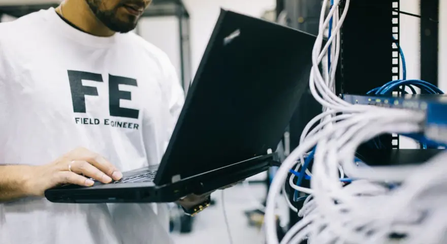 Inżynier w koszulce "FE FIELD ENGINEER" sprawdza sieć na laptopie. Analizuje połączenia, jakby sprawdzał, tracert co to jest.