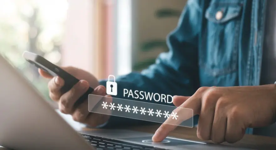 Osoba wpisuje hasło na laptopie, trzymając telefon. To dobry przykład, jak tworzyć silne hasło.